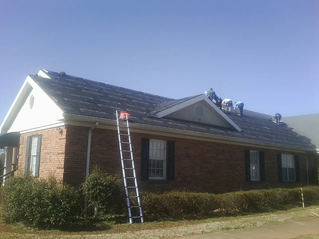 Crew tearing shingles off a brick ranch roof with a ladder set — Emergency Roof Repair work by our crew in Essex